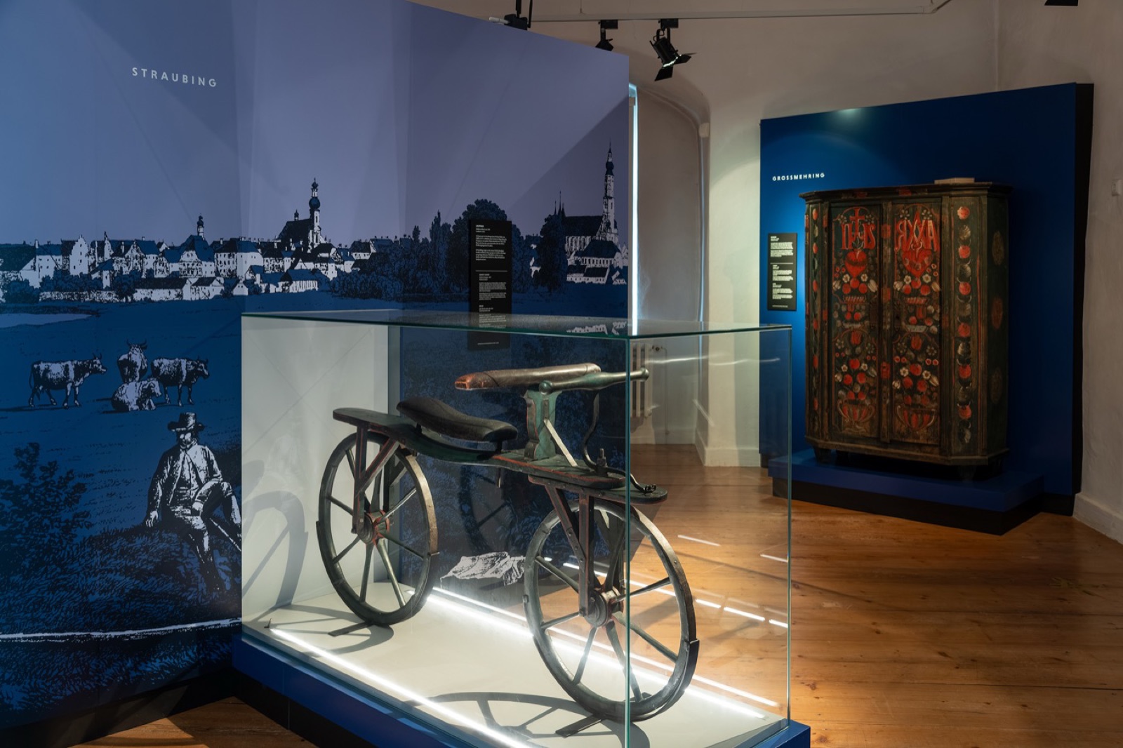 Ausstellungsraum mit einem hölzernen Laufrad von 1818 in der Vitrine, dahinter eine bemalte Truhe