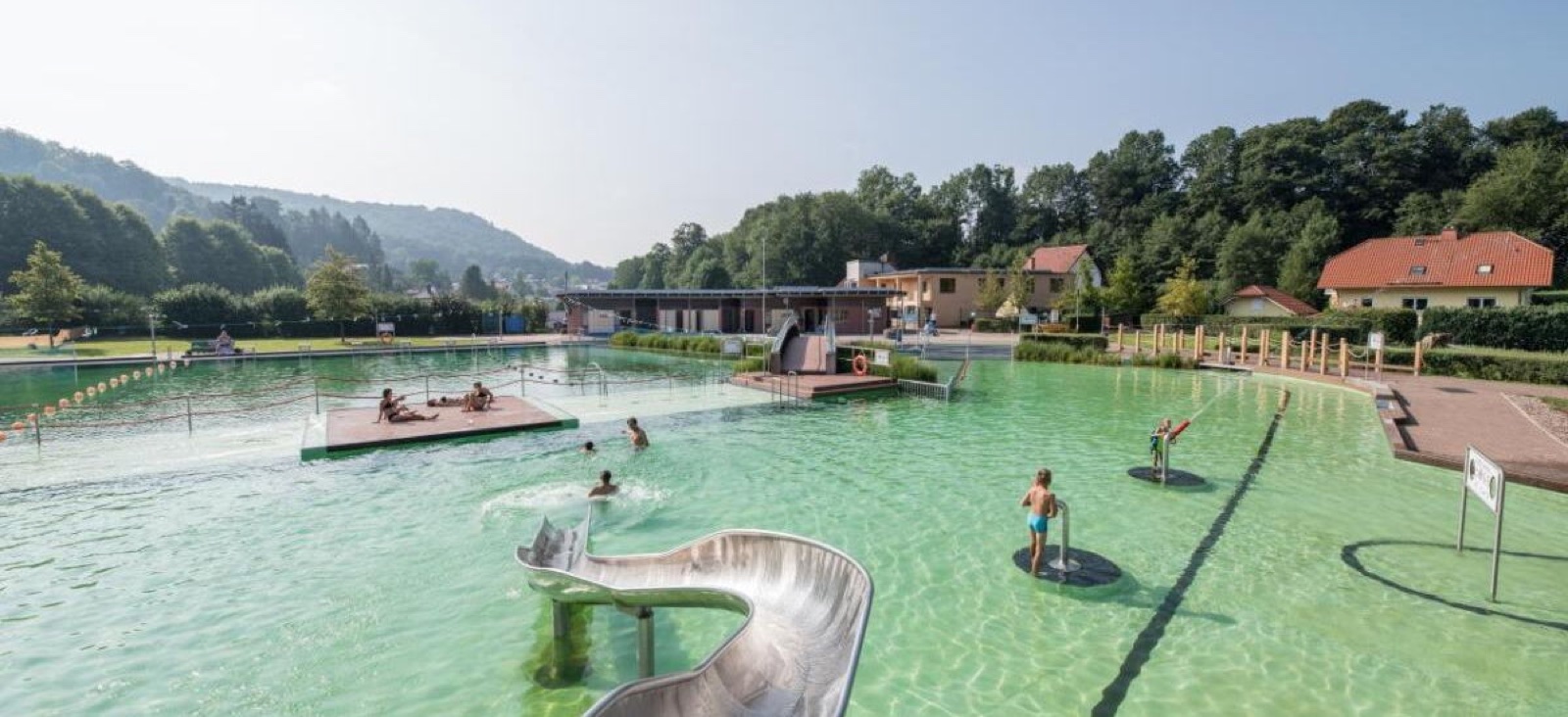 Blick über das Naturbad mit Rutsche, Schwimmbereich und bewaldeten Hügeln im Hintergrund