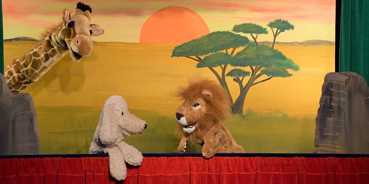 Giraffe, Hund und Löwe als Handpuppen auf der Bühne vor gemalter Savannenkulisse