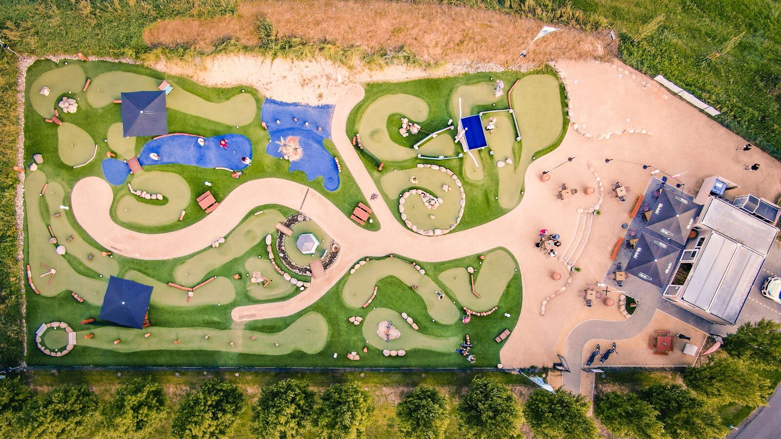 Luftbild der Adventuregolf-Anlage Family Fairway: 18 Bahnen mit Kunstrasen, blauen Wasserläufen und Findlingen, rechts der Coffee Shop