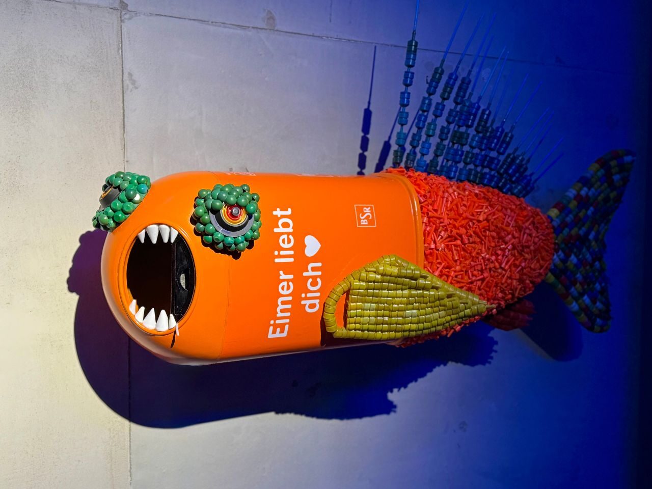 Fisch aus einer Mülltonne und buntem Plastikmüll vor blauer Wand